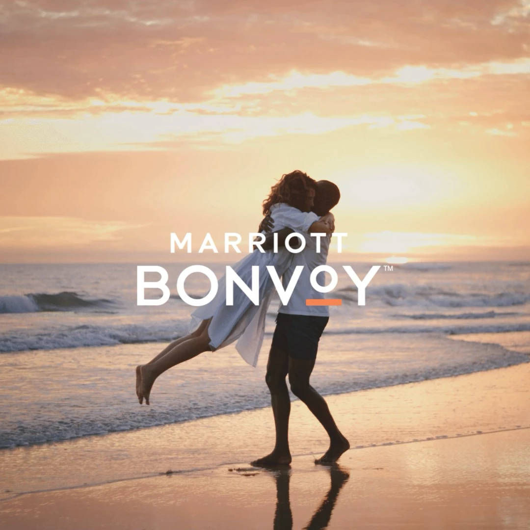 Couple embracing on a beach at sunset with Marriott Bonvoy logo overlay