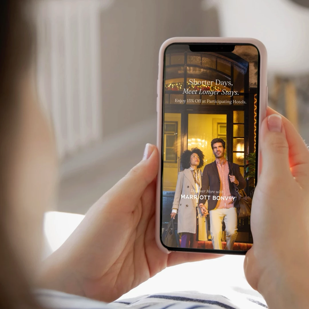 Couple standing at hotel entrance with Marriott Bonvoy promotional message on smartphone screen